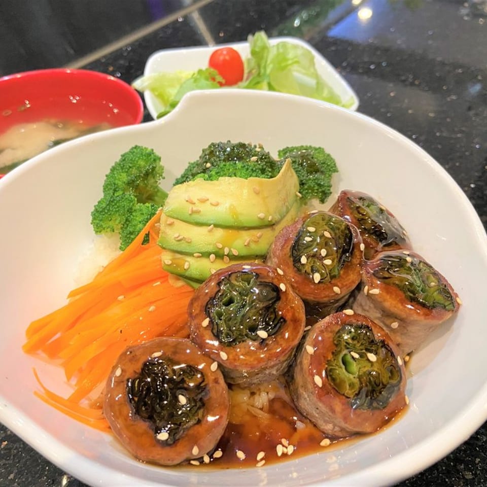 Japanese teriyaki bowl with beef rolls, broccoli, avocado, carrots, and sesame seeds, with soup and salad.